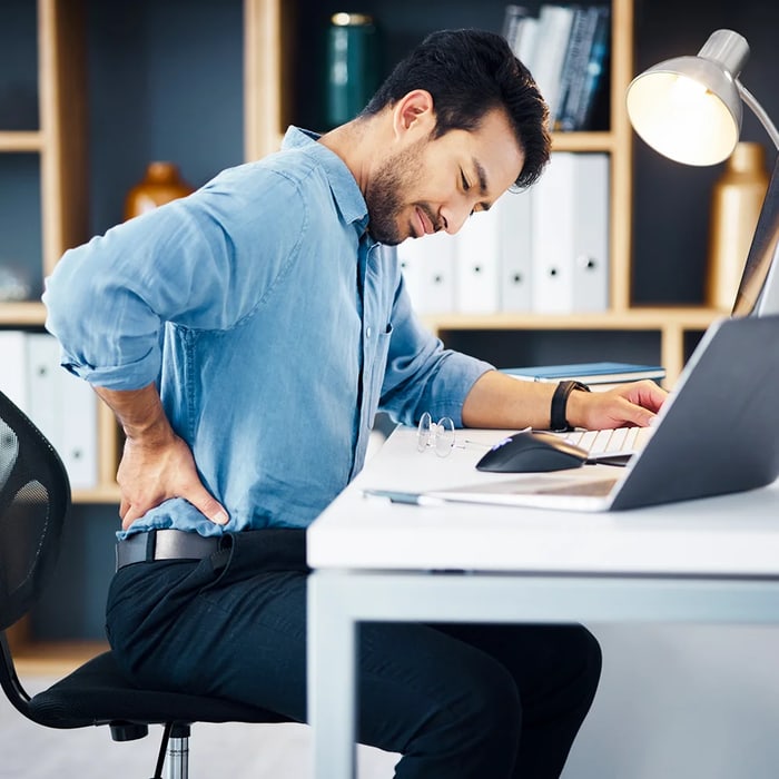 Man seated at a desk reaching for his aching lower back