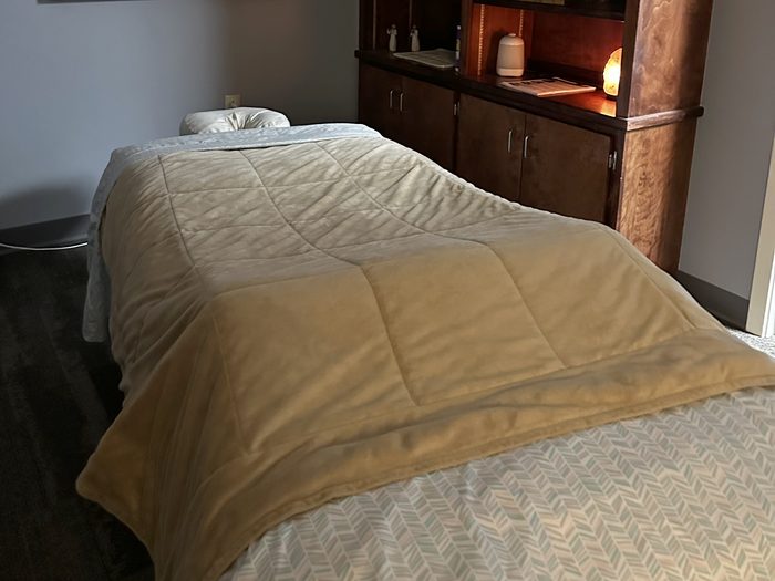 Massage table under a quilted blanket beside wood shelving and a warm salt lamp in the ProWellness massage therapy room